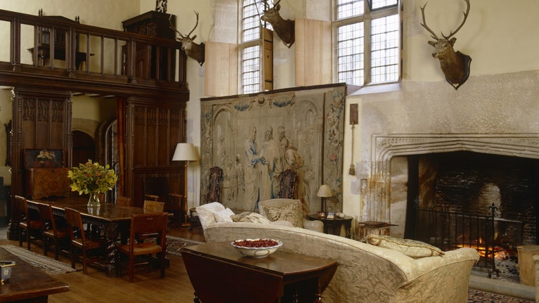 A view of the Great Hall at Great Chalfield laid out as a sitting room, with a tapestry and deer heads on the wall, a sofa in front of the lit fire and a dining table.